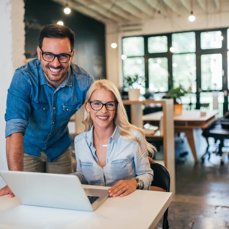 Two smiling professionals in a modern office collaborate at a laptop, showcasing teamwork and innovation.