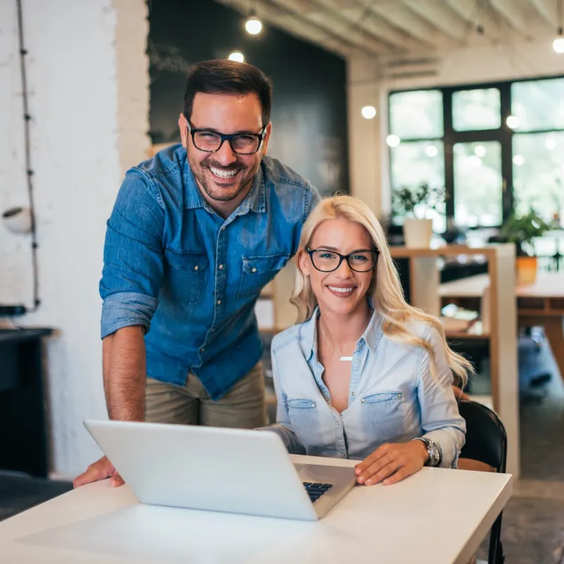 Smiling man and woman wearing glasses working together on a laptop in a modern office space.