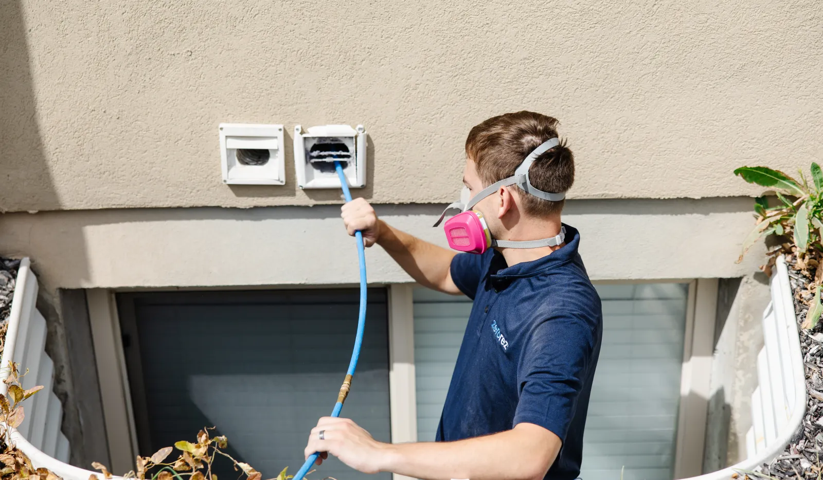 Technician wearing a respirator mask cleaning dryer vents on a residential building using a blue hose brush.