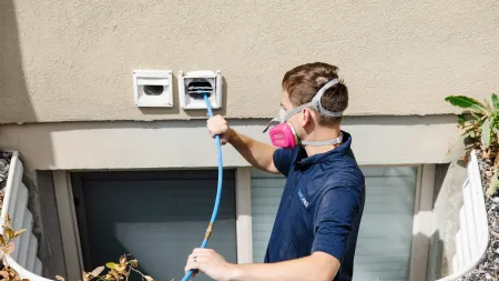 Technician wearing a respirator mask cleans exterior dryer vents with a blue brush attached to a hose.