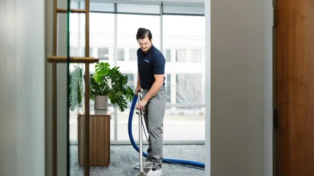 Professional cleaner using carpet extractor machine in modern office hallway with plants and large windows.
