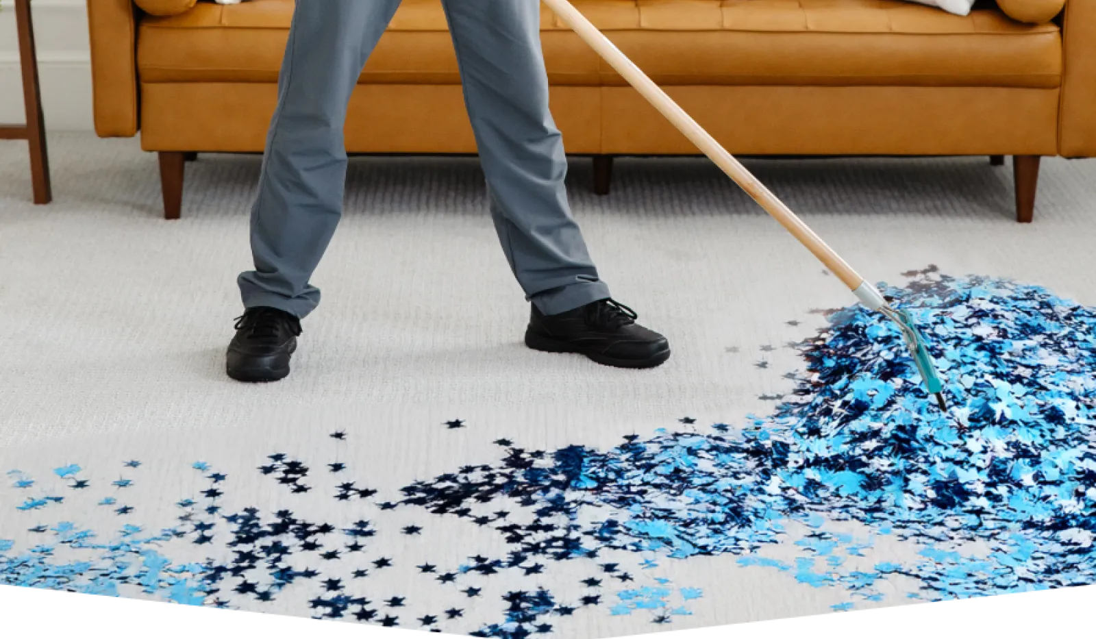 Person sweeping blue and black star-shaped confetti on beige carpet in living room with brown sofa.