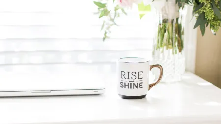 White desk with a laptop, Rise and Shine mug, and vase of pink and white flowers by a sunlit window.
